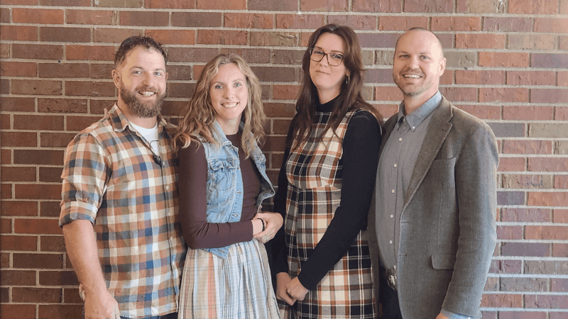 A photo of four adults standing in front of a brick wall.