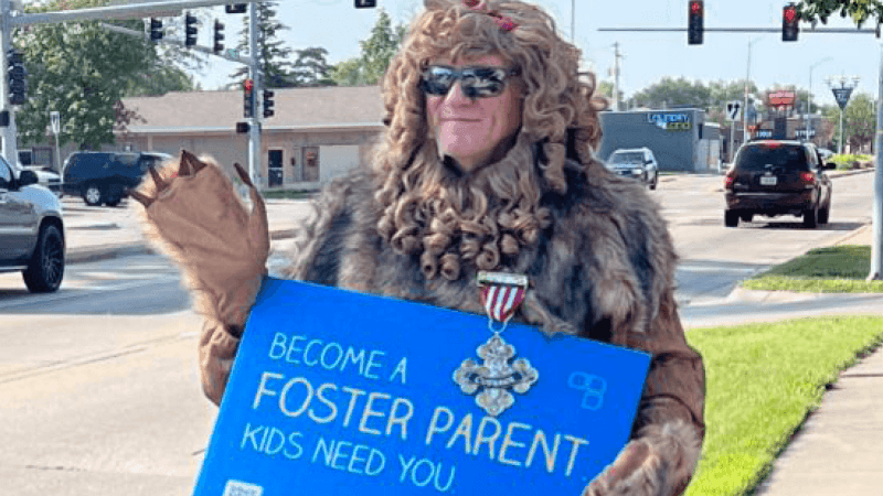 Pete in a lion costume holding a 'become a foster parent' sign and waving to traffic