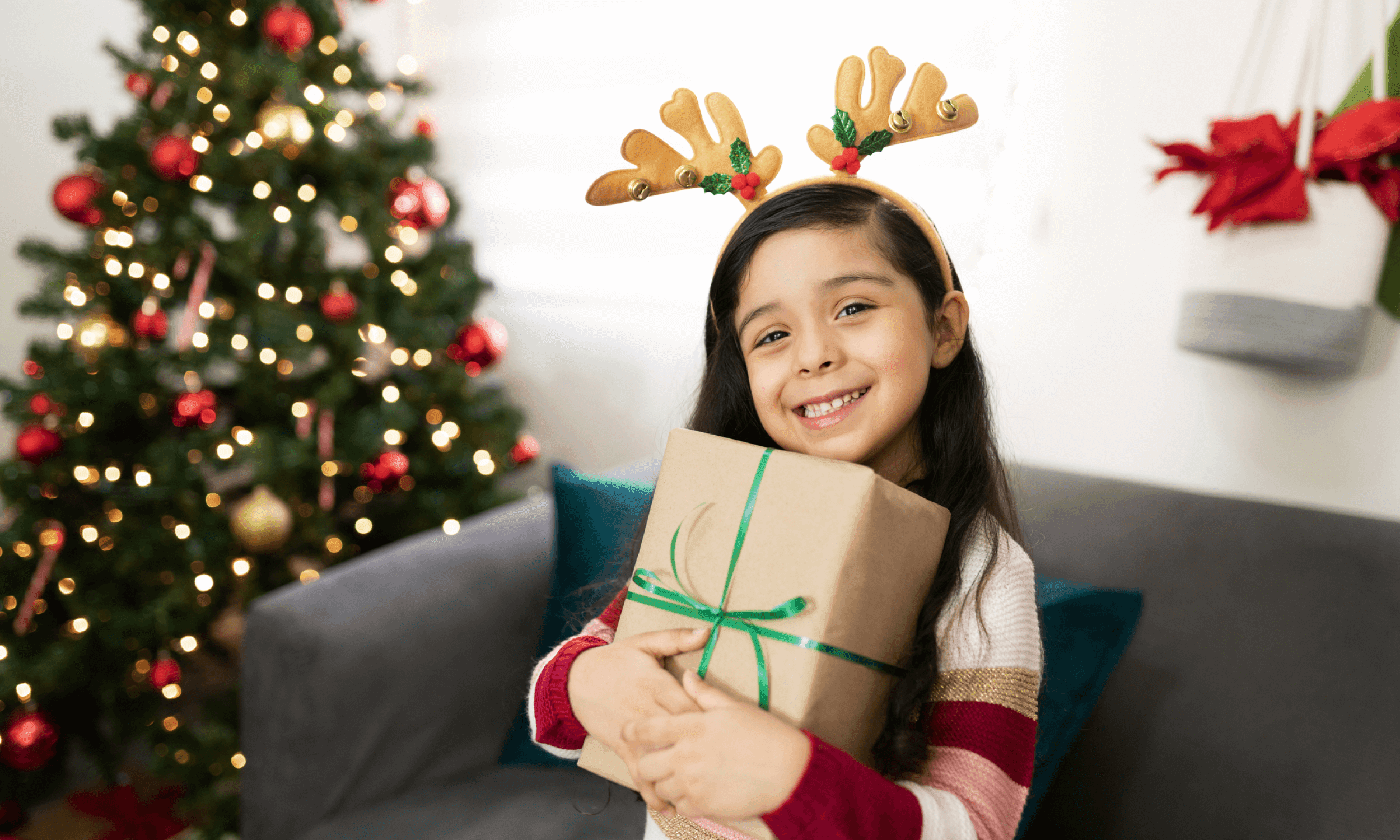 School-aged girl wearing antlers and holding a gift next to a Christmas tree